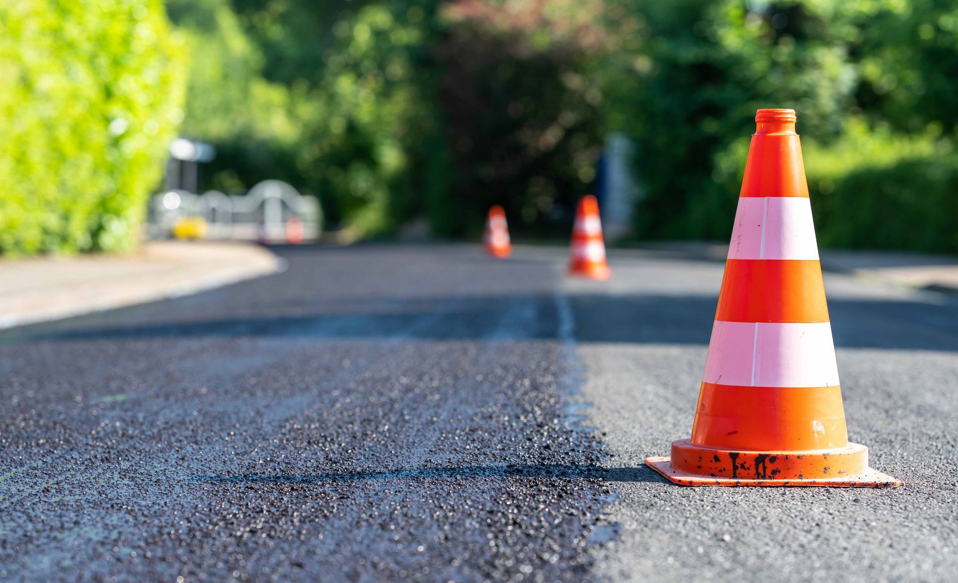 Orange and white traffic cones stand on a freshly paved asphalt road with green trees in the background.