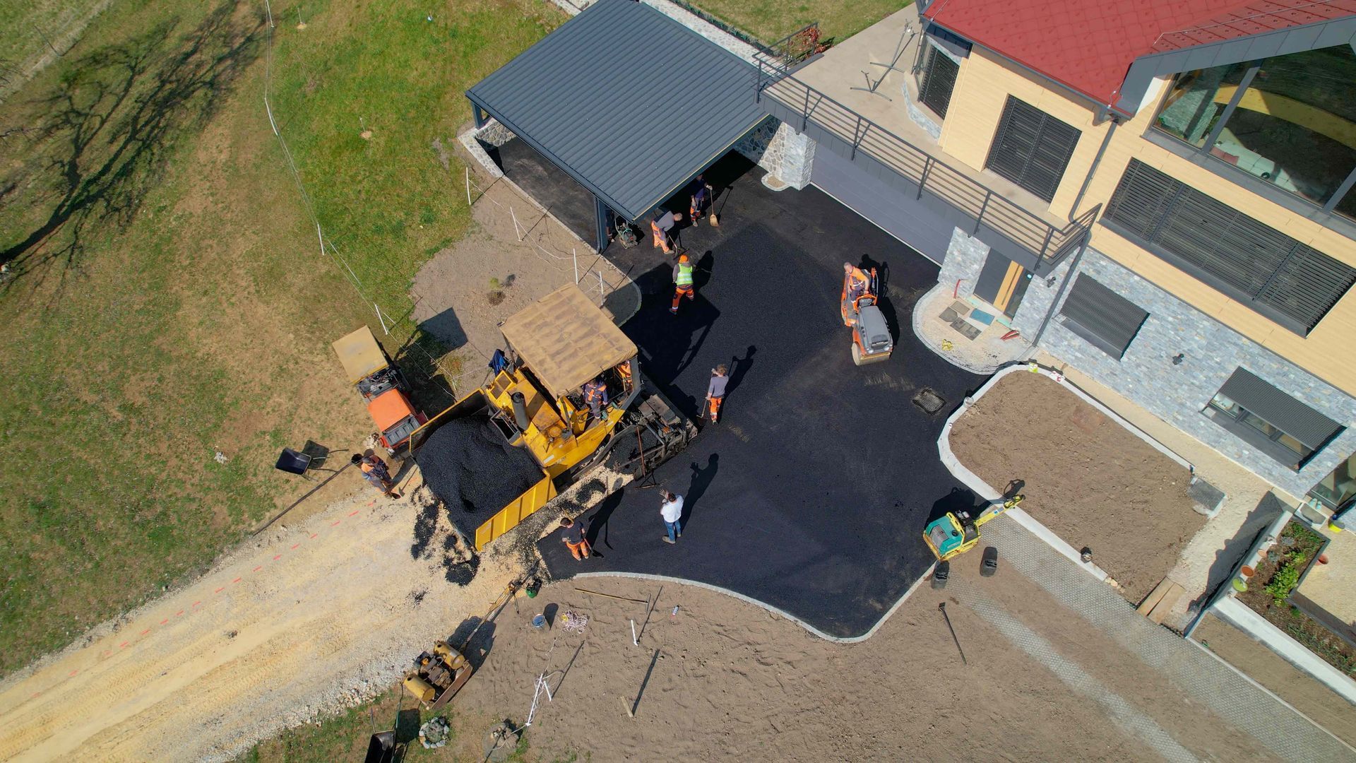 Aerial view of construction workers paving a residential driveway with machinery near a house on a sunny day.