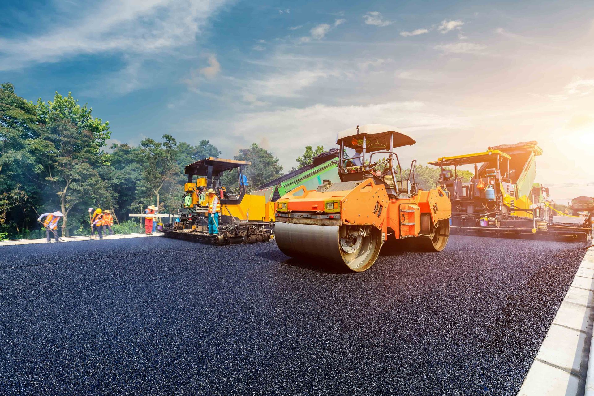 Construction workers operate yellow and orange road rollers to pave a new asphalt road at sunset.