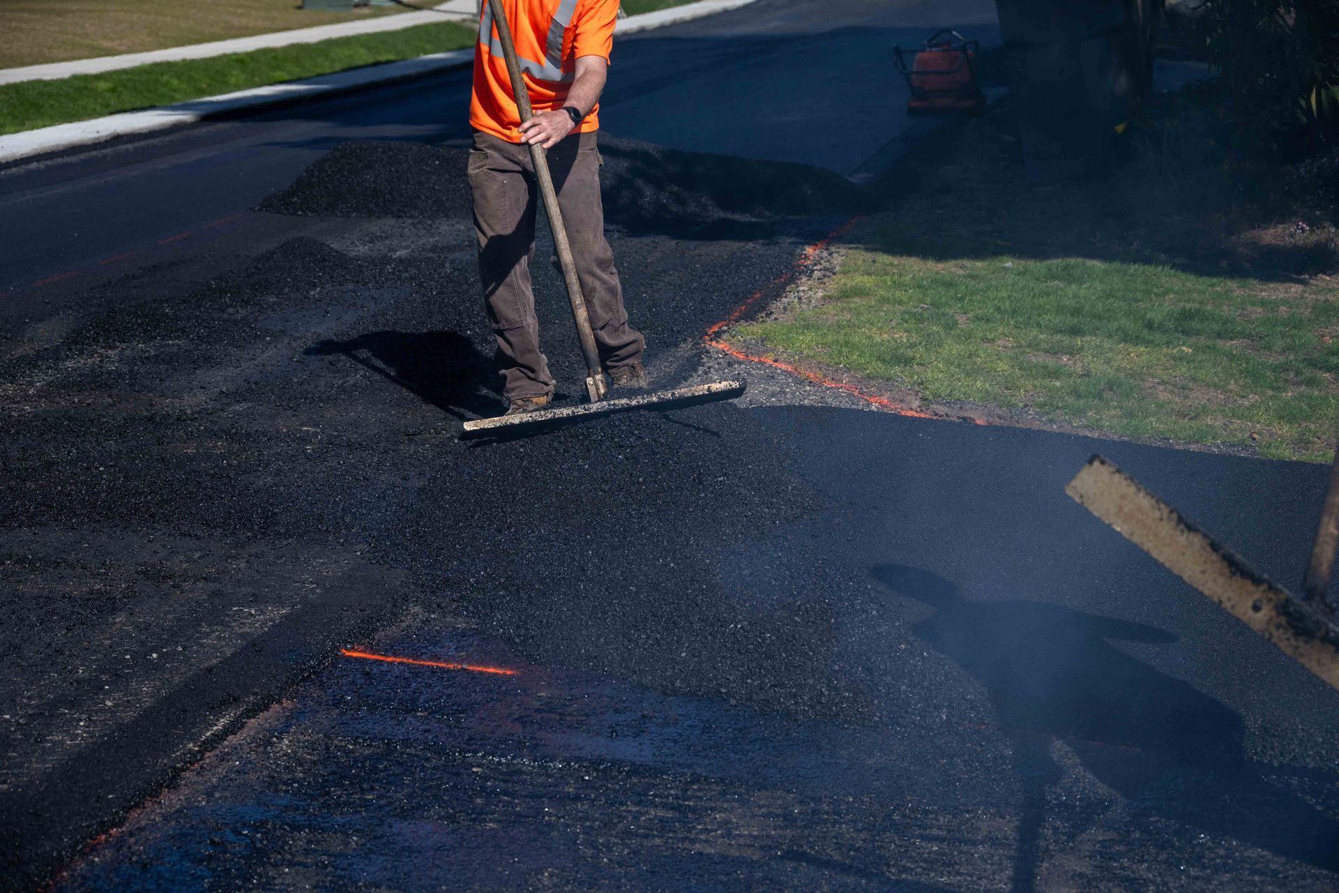 A construction worker in an orange safety vest uses a rake to spread fresh black asphalt on a paved road.