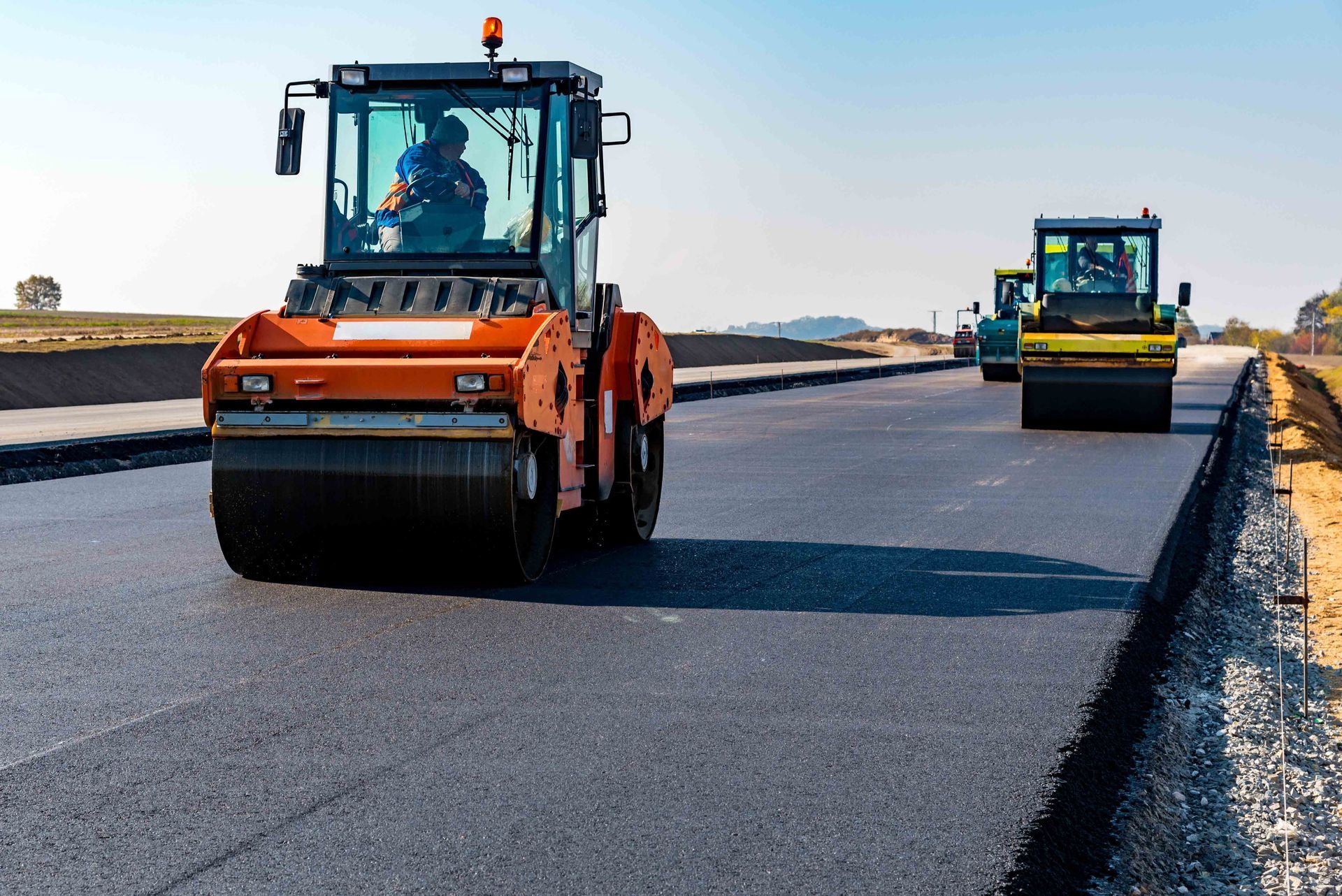 Two orange and yellow road rollers compact fresh black asphalt on a sunny highway construction site.