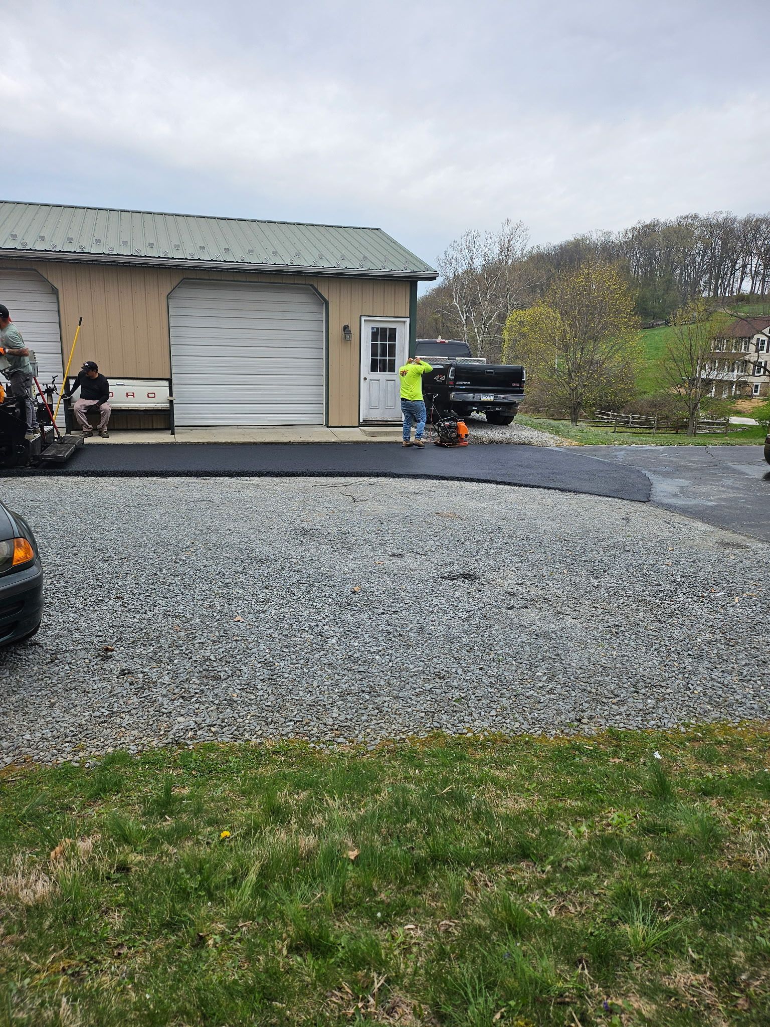 A person in work clothes spreads fresh asphalt next to a house while a yellow steamroller compacts it nearby.