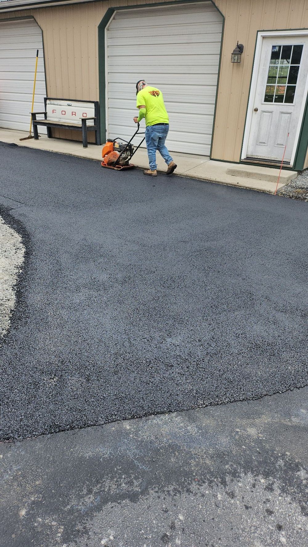 A worker in an orange safety vest smooths freshly laid black asphalt on a road near a steamroller. A worker in an orange safety vest smooths freshly laid black asphalt on a road near a steamroller.