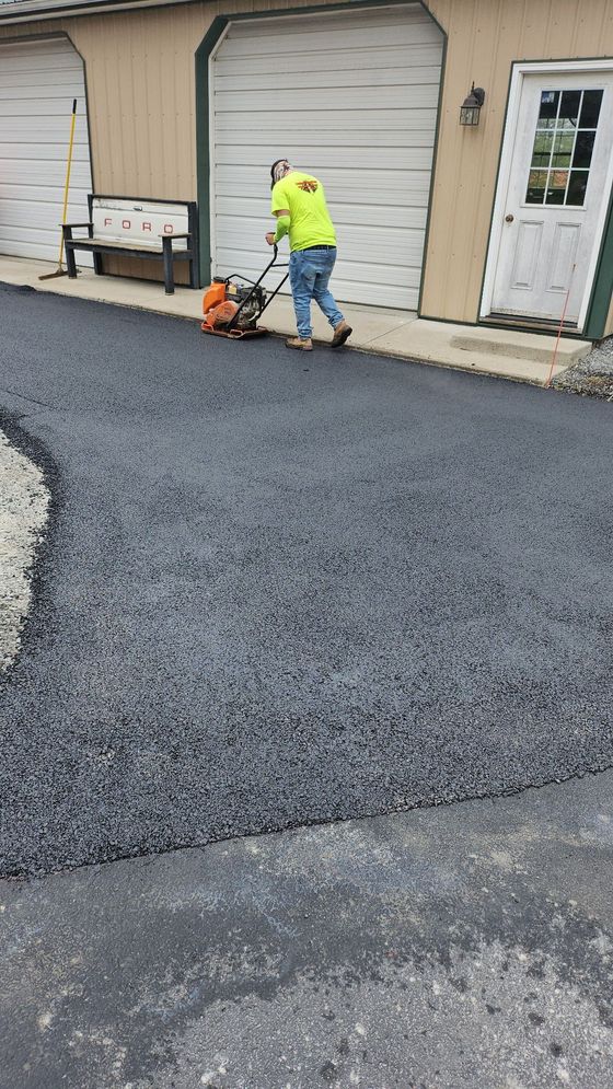 A worker in an orange safety vest smooths freshly laid black asphalt on a road near a steamroller.
