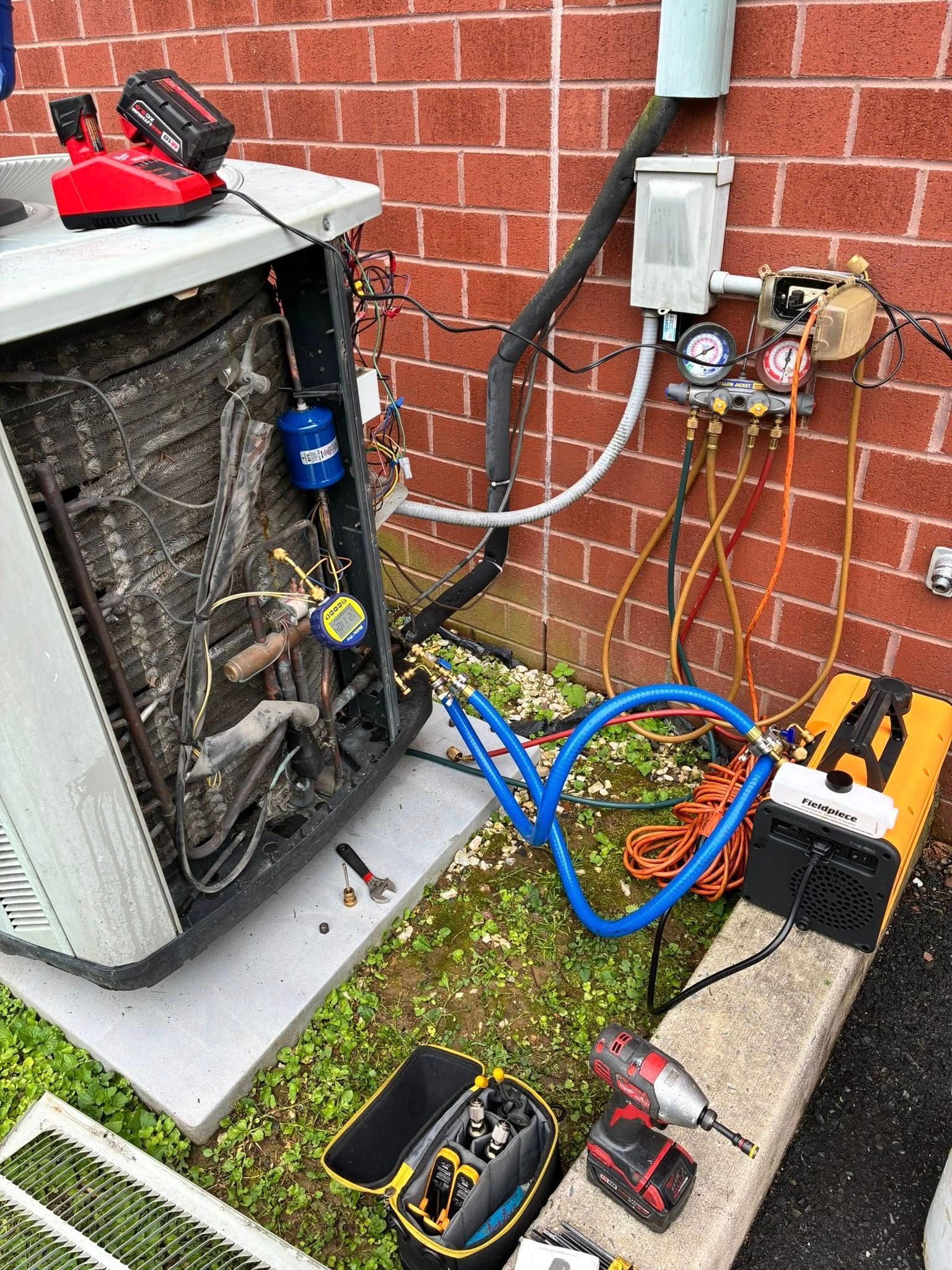 An outdoor HVAC unit under repair, with gauges, vacuum pump, and tools connected to the unit against a brick wall.