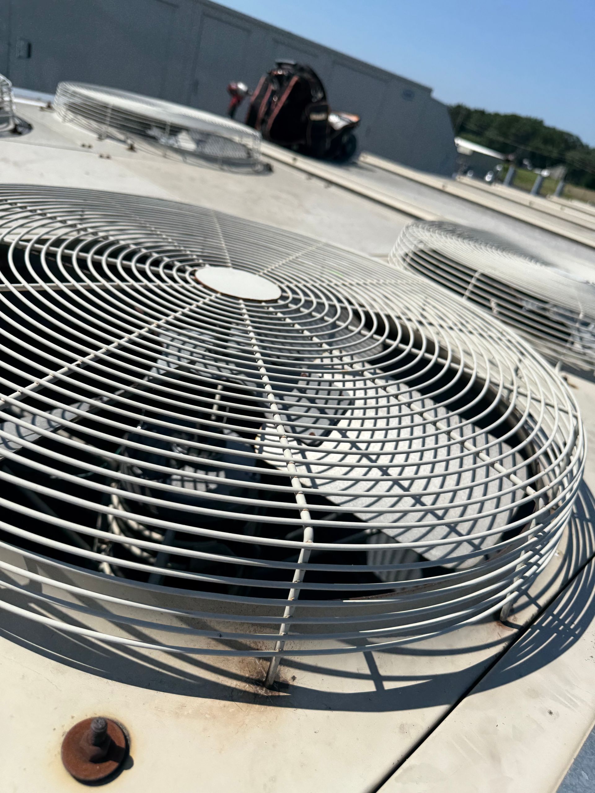 Close-up of a metal circular fan guard on a rooftop HVAC unit with more units in the background under a blue sky.