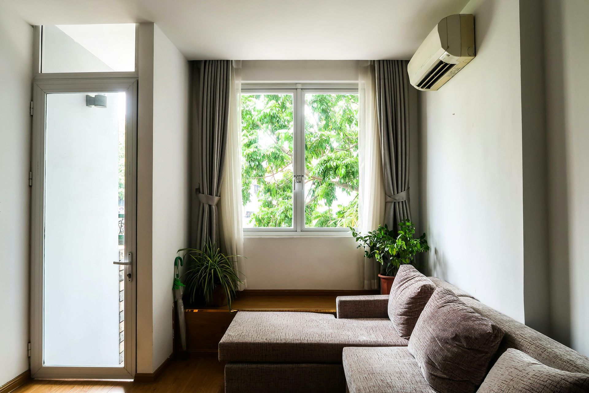 A modern living room with a gray sectional sofa, a large window with greenery outside, and a wall-mounted air conditioner.