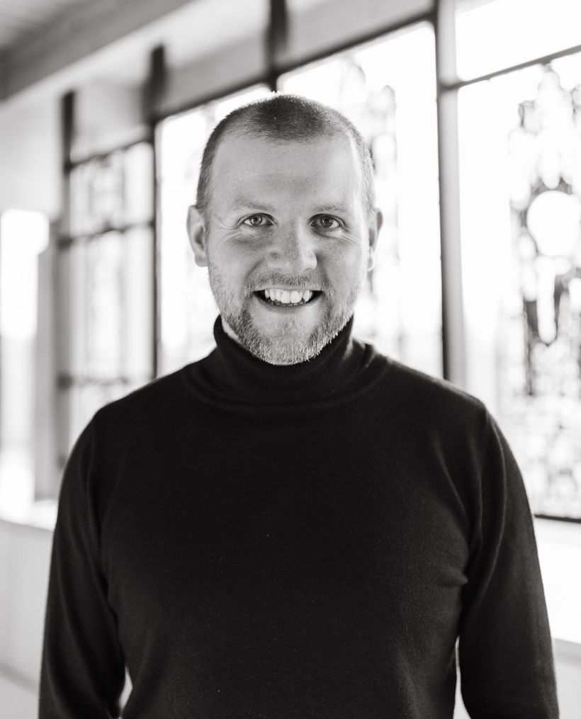 A man with a beard is wearing a black turtleneck sweater and smiling in a black and white photo.