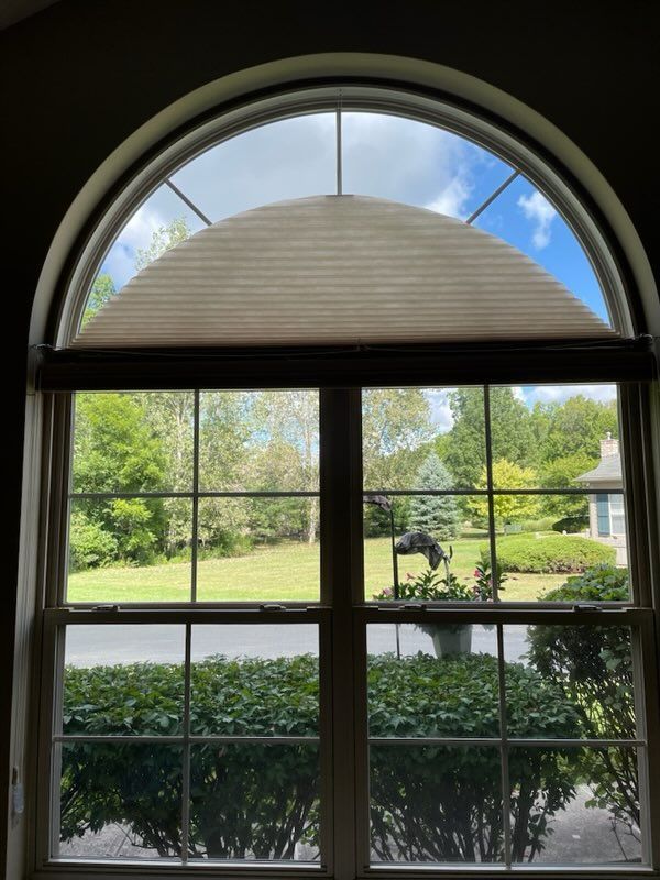 Window with arched top, view of green yard and blue sky. Beige shade partially covers arched pane.