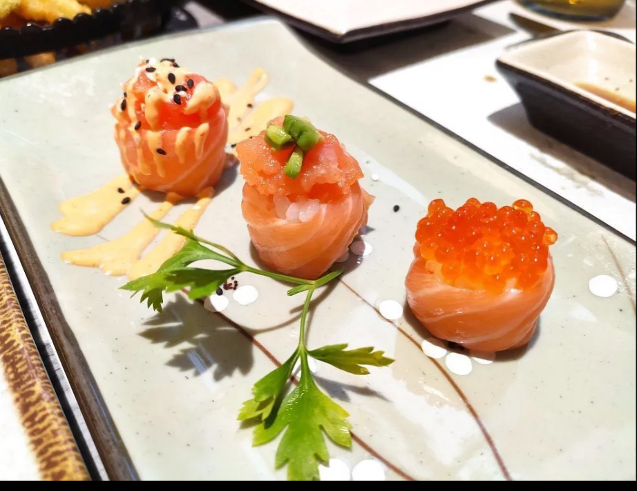 Tre bocconcini di sushi al salmone, guarniti e disposti su un piatto bianco a un tavolo di ristorante.