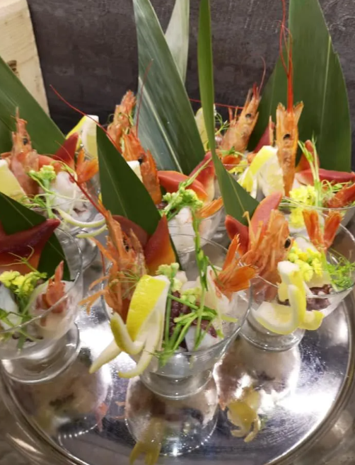 Antipasti di gamberi guarniti in piccoli bicchieri su un vassoio riflettente con decorazioni di foglie verdi.
