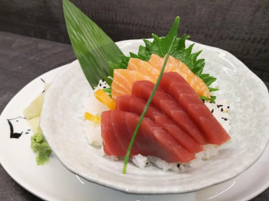 Piatto di sushi con sashimi di tonno, fette di salmone, riso e guarnizioni su un piatto bianco.