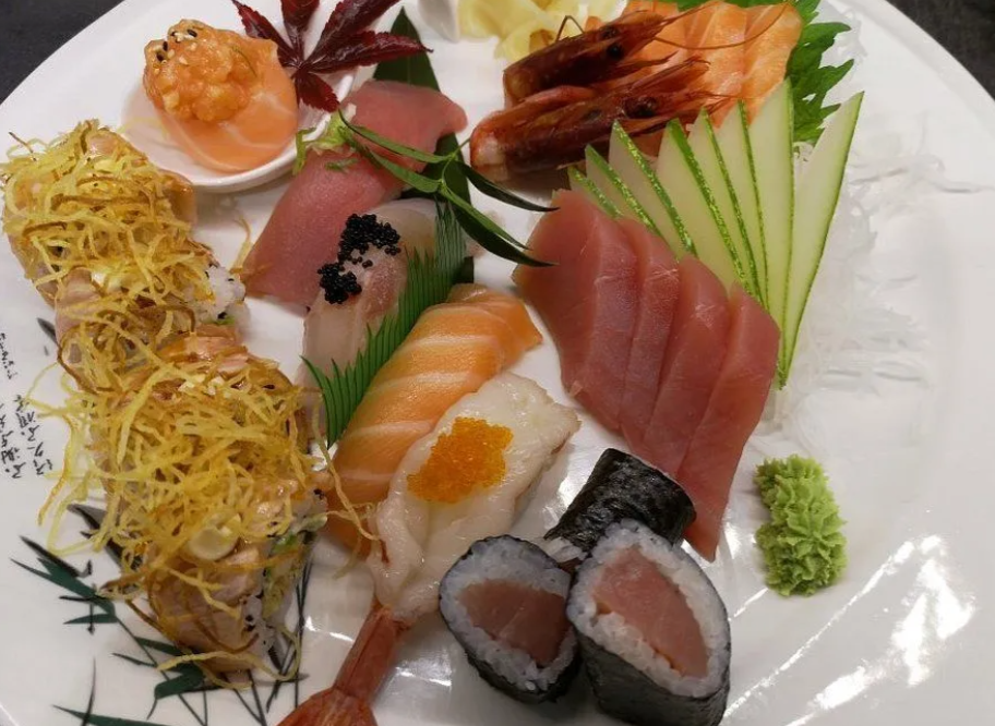 Piatto misto di sushi con sashimi, roll, gamberi e guarnizioni su un piatto bianco.