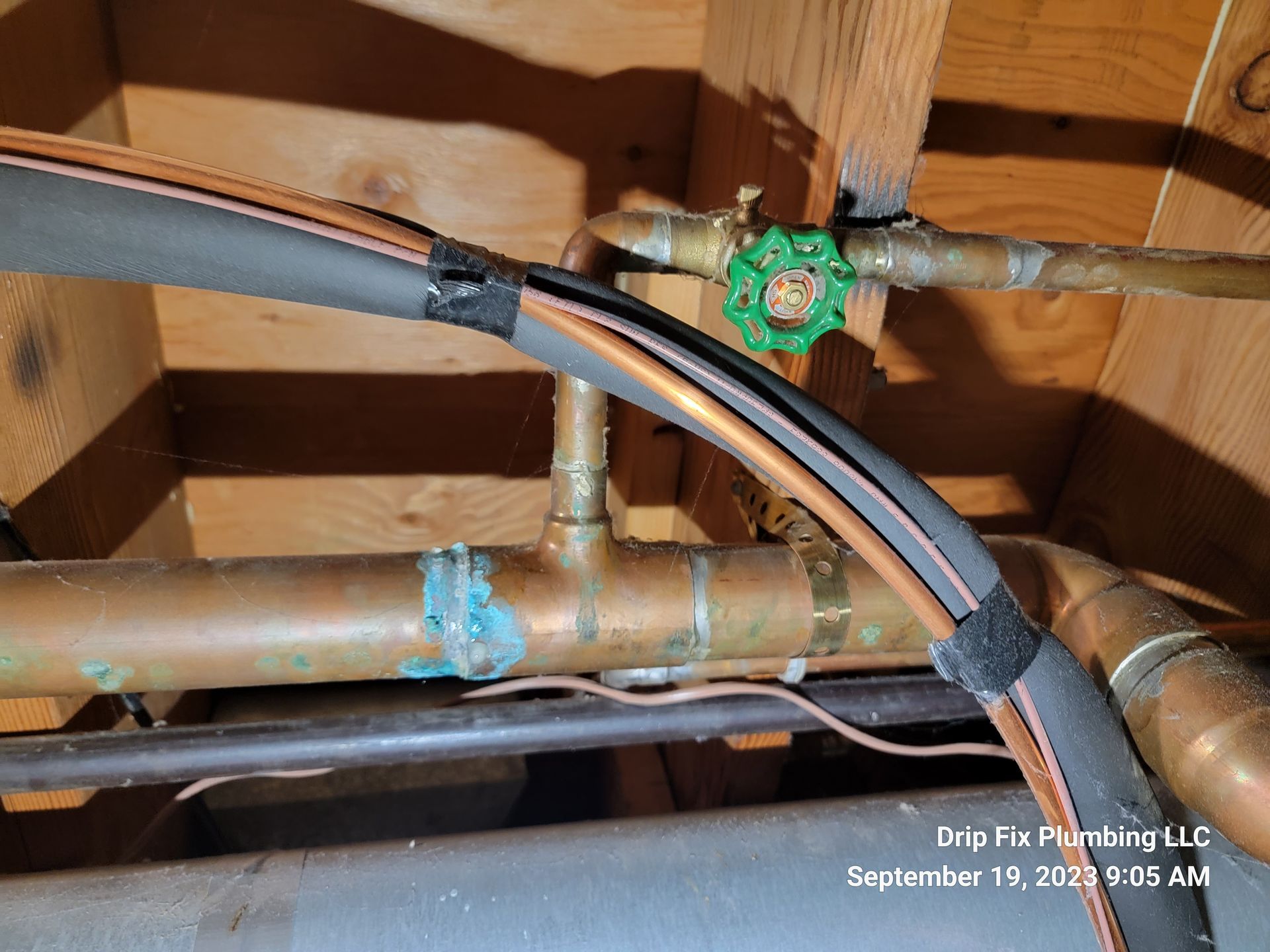 Copper pipes with green-handled valve, black insulation, and signs of corrosion in a wooden setting.