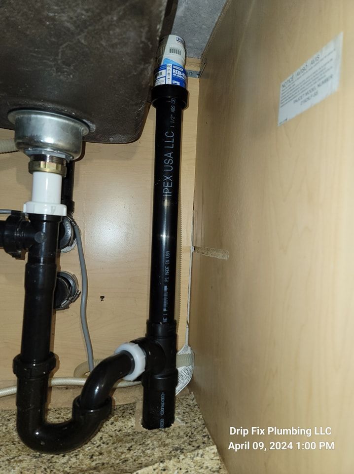 Black plumbing under a kitchen sink. The P-trap is visible, connecting to a drain pipe. Beige cabinets surround.