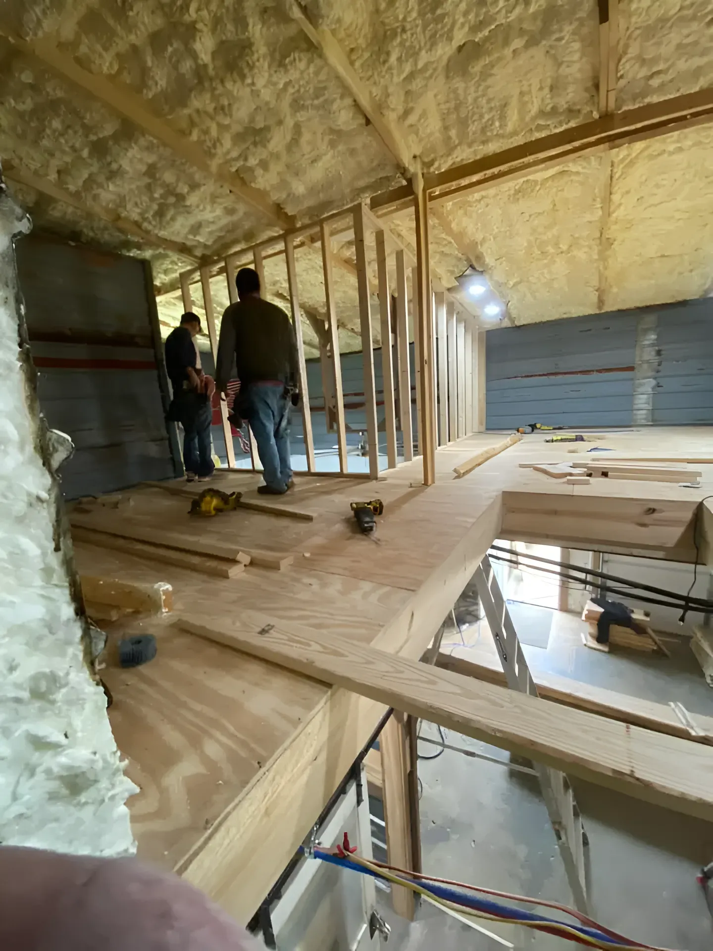 Construction site with two workers framing a wall in an attic space. Brown wood, white insulation visible.