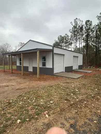 White and gray metal building with a porch, two garage doors, and concrete pads, set in a yard with trees.