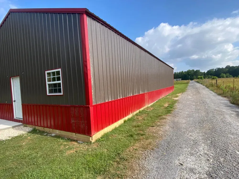 A large, brown and red metal building next to a gravel road, with a blue sky backdrop.