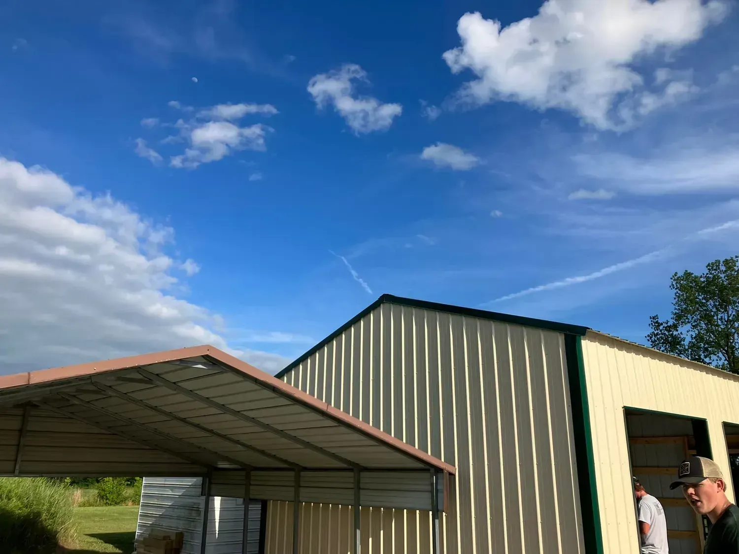 Carport and building with tan metal siding against a blue sky with clouds. Two men are visible.