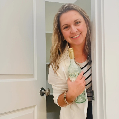 Woman in a white cardigan holding a green wine bottle, peeking out of a white closet.