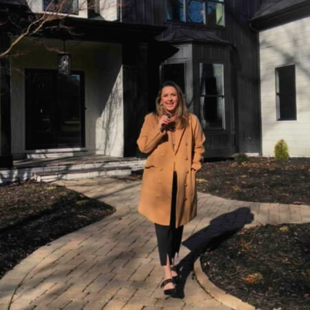 Woman in tan coat stands on a brick path in front of a dark house, smiling.