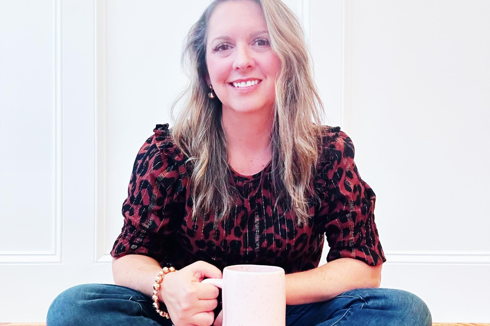 Woman with blonde hair, smiling, holding a pink mug, sitting cross-legged in front of a white wall.