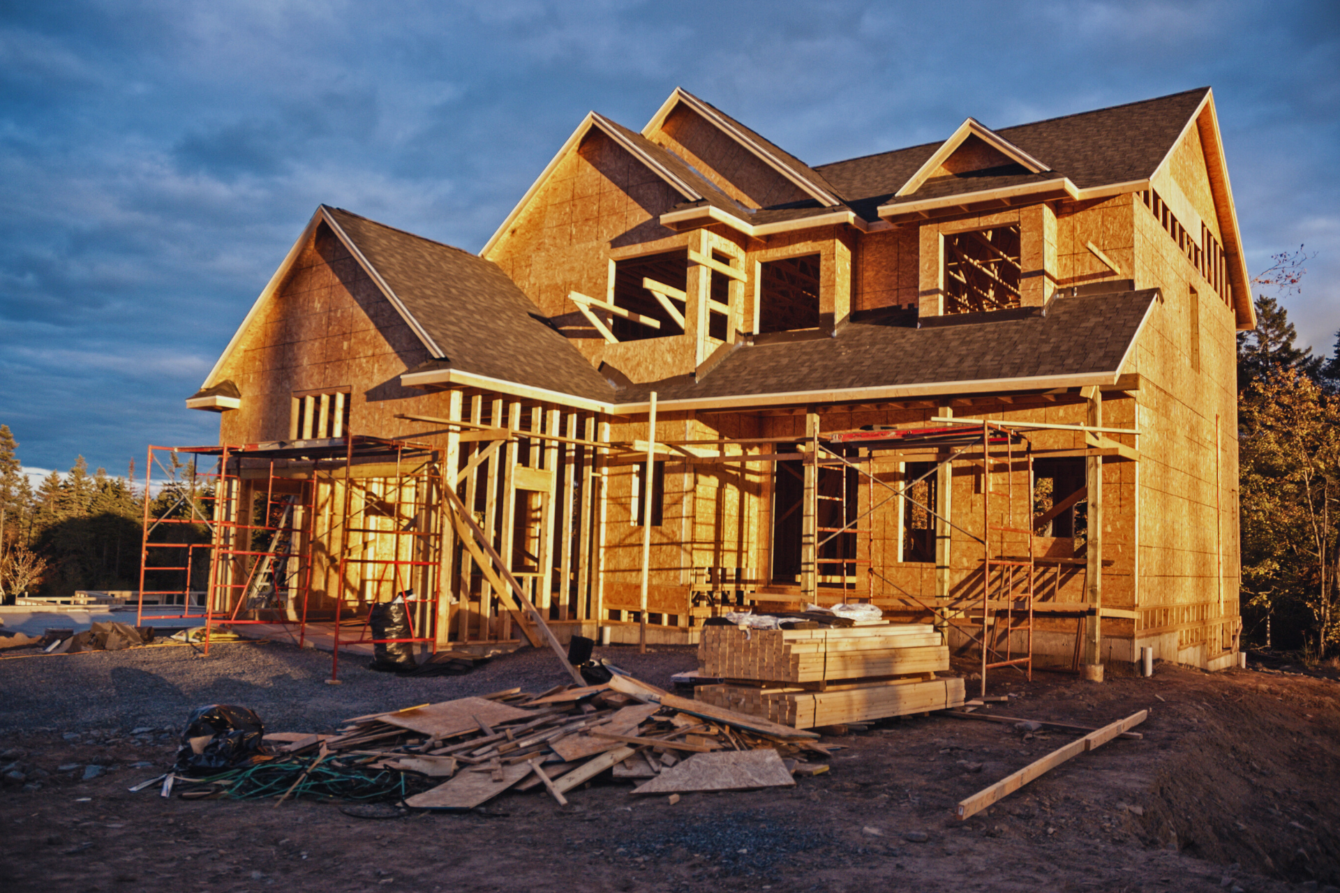 House under construction with exposed wooden frame. Brown siding, blue sky. Building materials on the ground.