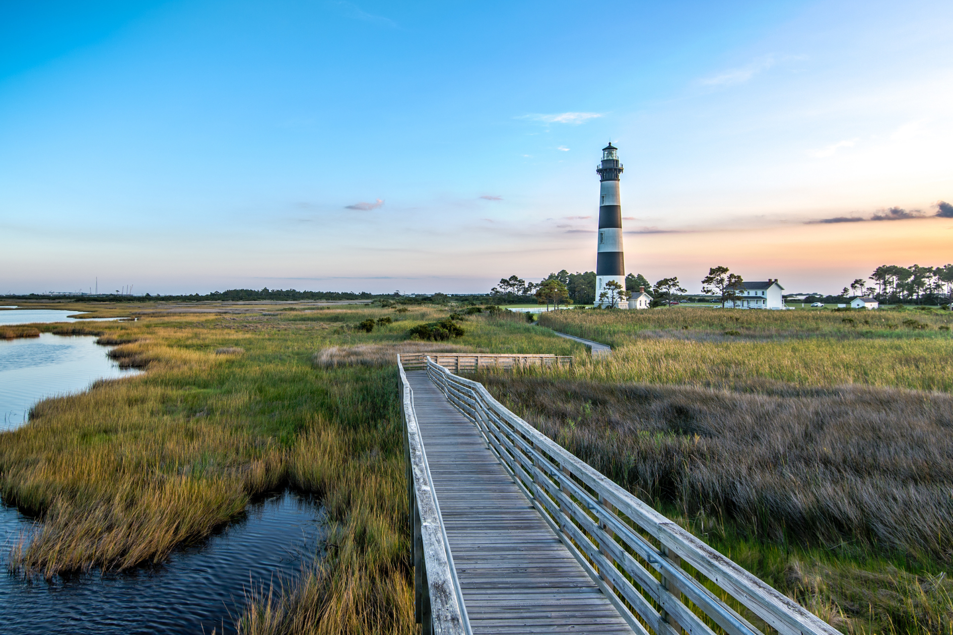 Wooden boardwalk leading to a black and white striped lighthouse by marshland at sunset.