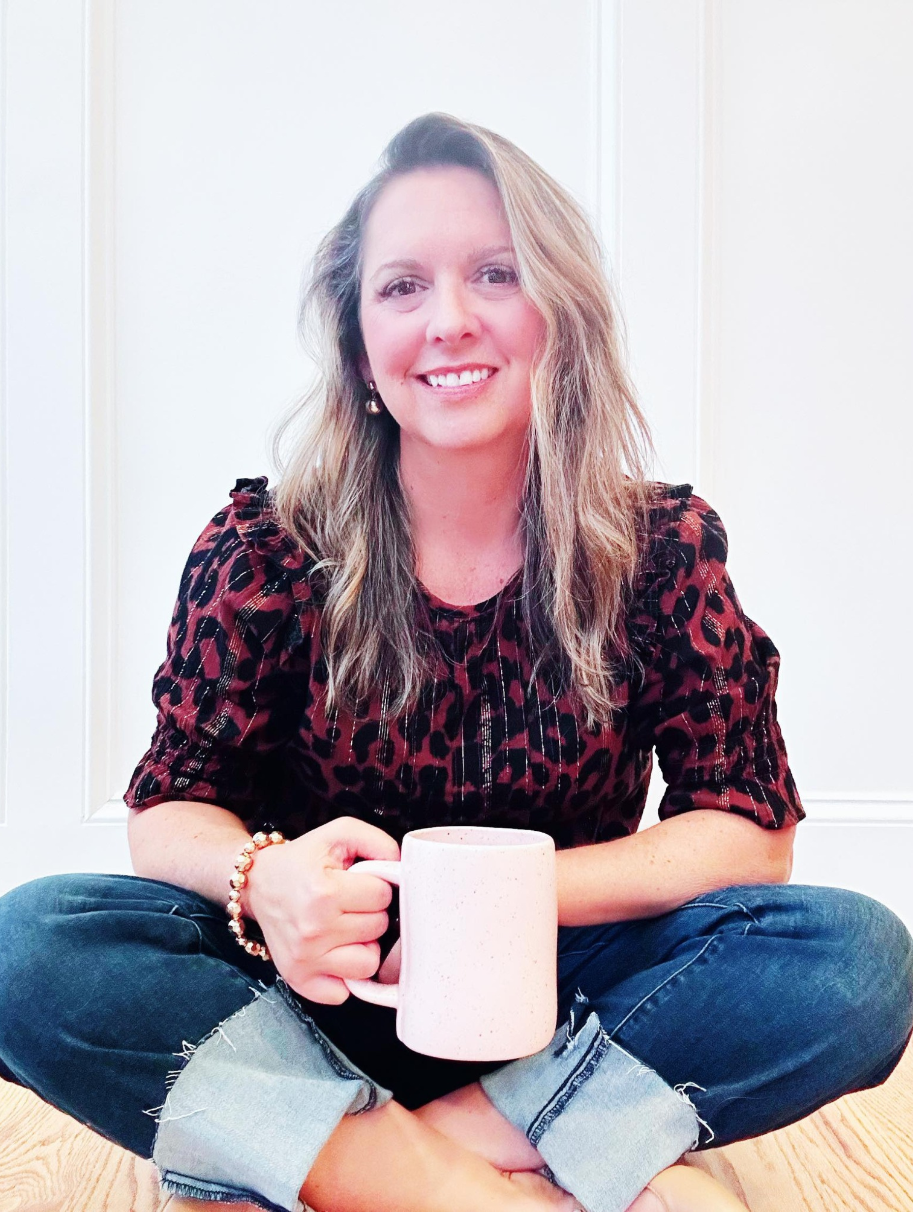 Woman seated cross-legged on floor, holding a mug. Smiling, with long hair, wearing jeans and patterned top.