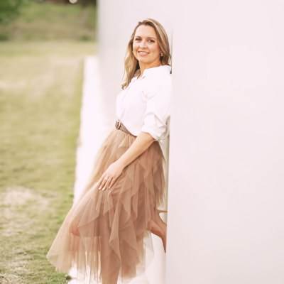 Woman in white shirt and brown tutu skirt leans against a white wall, smiling.