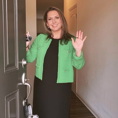 Woman in a green jacket waves from a doorway, holding keys. She smiles.