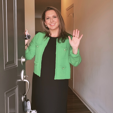 Woman in a green jacket waves from a doorway, holding keys. She smiles.