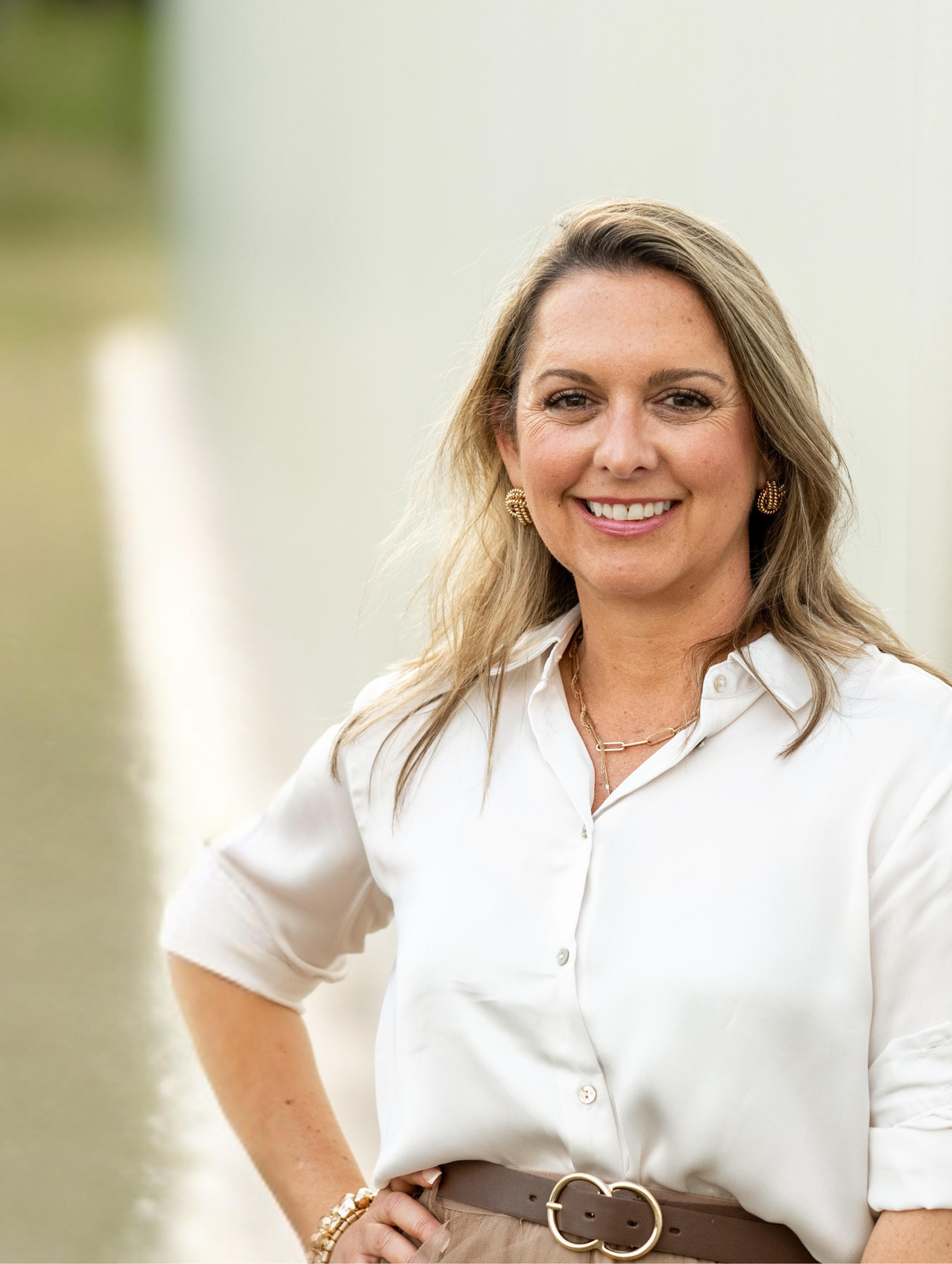 Woman smiling, hand on hip, wearing white blouse, gold jewelry, standing outdoors.