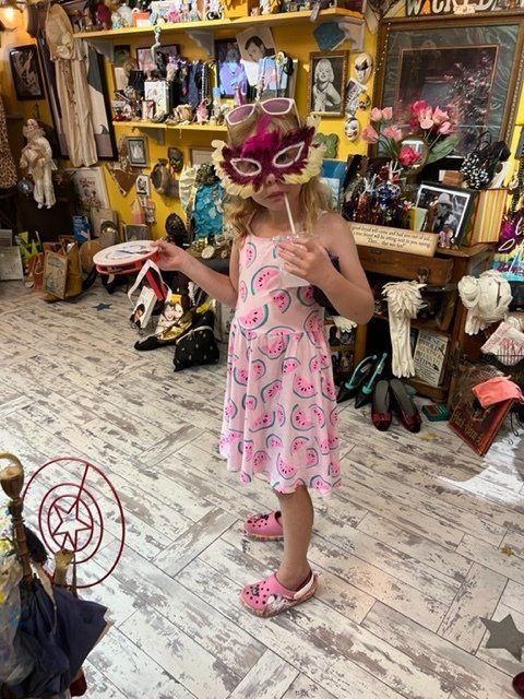 A little girl wearing a mask and sunglasses is holding a lollipop.