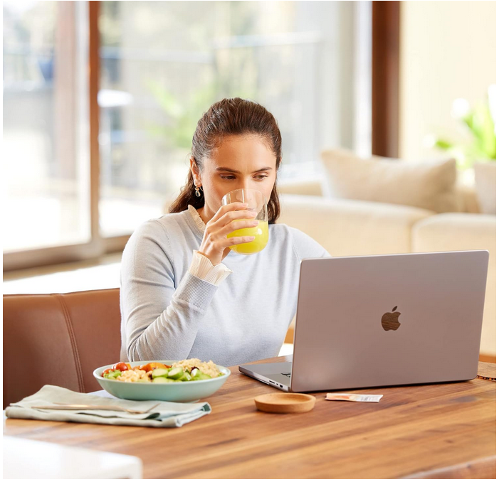 A woman is sitting at a table drinking orange juice and looking at a laptop.