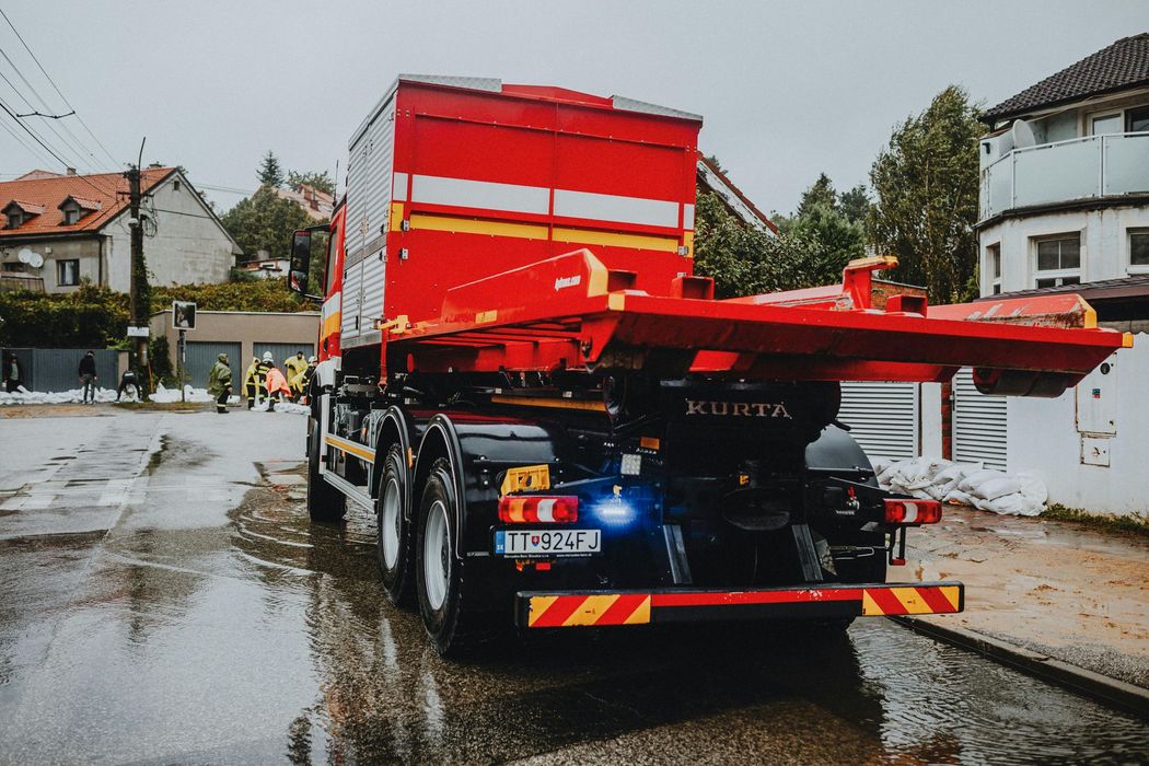 Red truck with attached container parked on a wet street; houses and people in the background.
