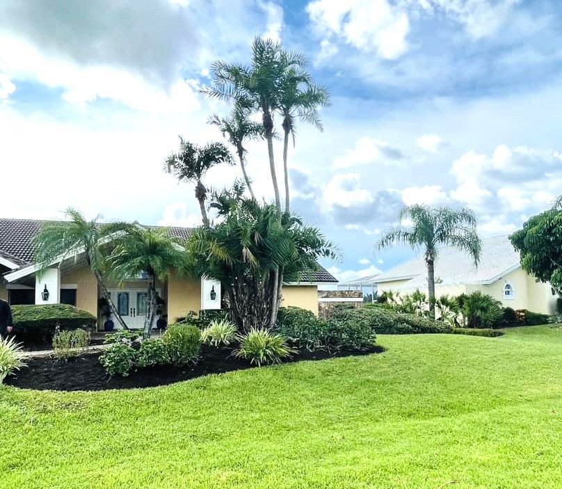 A yellow house with lush landscaping, palm trees, and a green lawn under a cloudy sky.