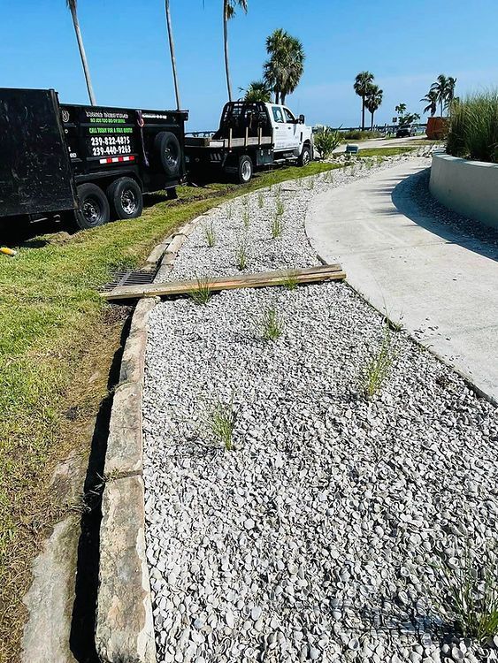 A gravel-covered landscaped area with small green plants alongside a curved sidewalk. A truck and trailer are parked nearby.