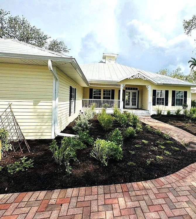 Yellow house with a metal roof, a landscaped garden, and a brick pathway leading to the entrance. Sunny day.