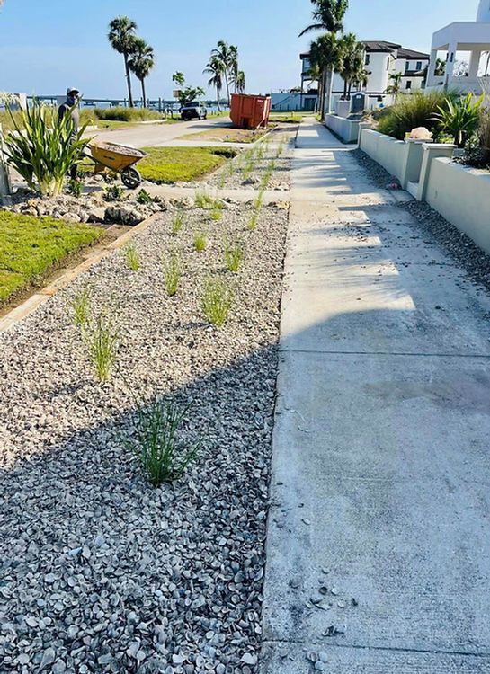 A paved sidewalk with a gravel and plant bed on one side, leading towards a beach. Sunny day.