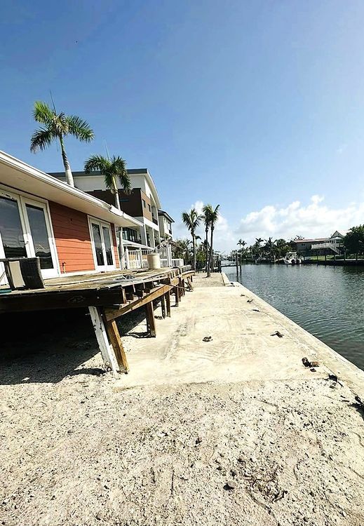 A waterfront property with a deck, concrete dock, and a canal under a bright blue sky with palm trees.