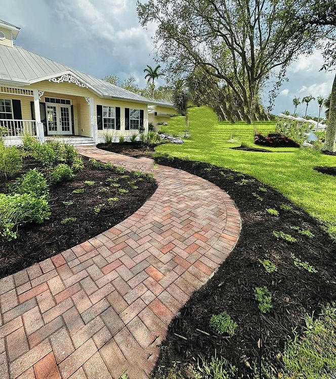 Brick pathway curves toward a yellow house with a white porch, through freshly mulched flower beds and a grassy lawn.