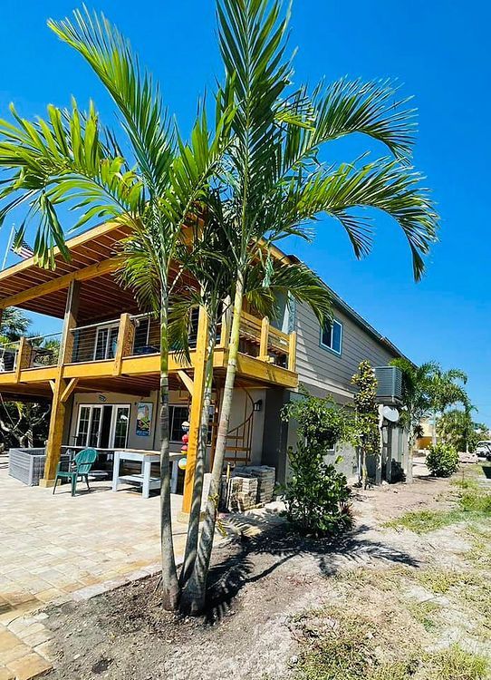 A two-story house with a wooden deck and palm trees against a blue sky. There's a paved patio in front.