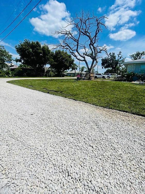 A gravel driveway leads past green grass and a leafless tree under a blue sky. In the background, are a few houses.