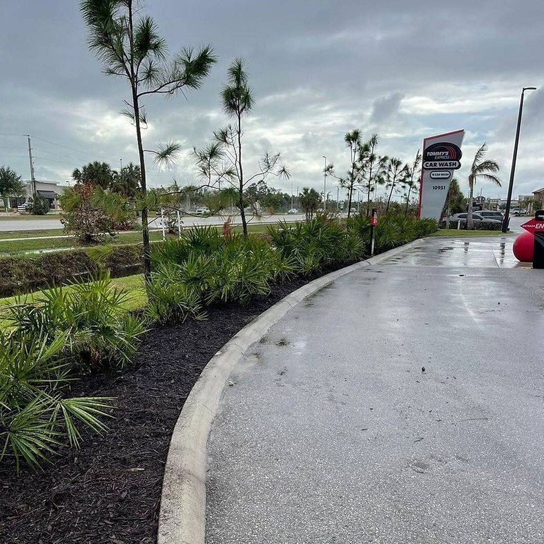 A wet, paved area with a landscaped border of plants and mulch in front of a commercial sign under a cloudy sky.
