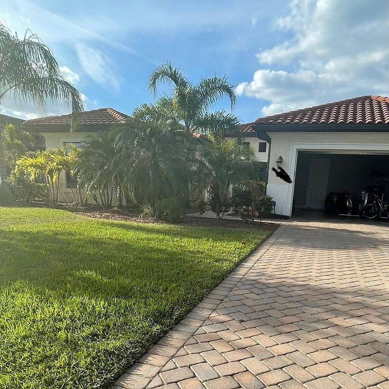 A one-story house with a red-tiled roof, palm trees, and a paved driveway on a sunny day. The garage door is open.