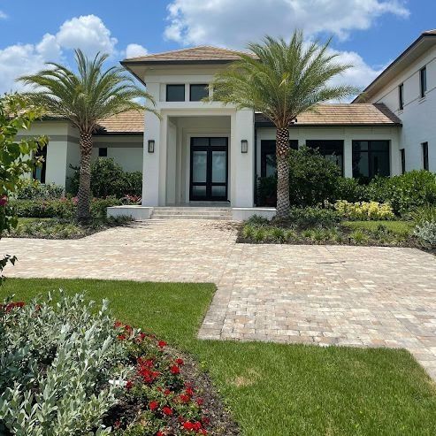 A light-colored, modern house with palm trees and a brick driveway on a sunny day. Landscaped gardens surround the entrance.