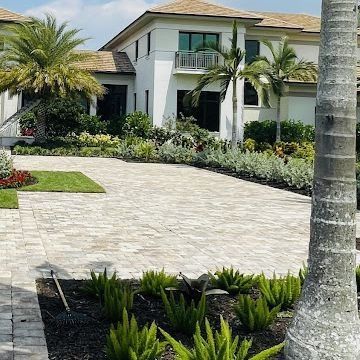 A large white house with a tiled driveway and tropical landscaping. Palm trees and green plants surround the home.
