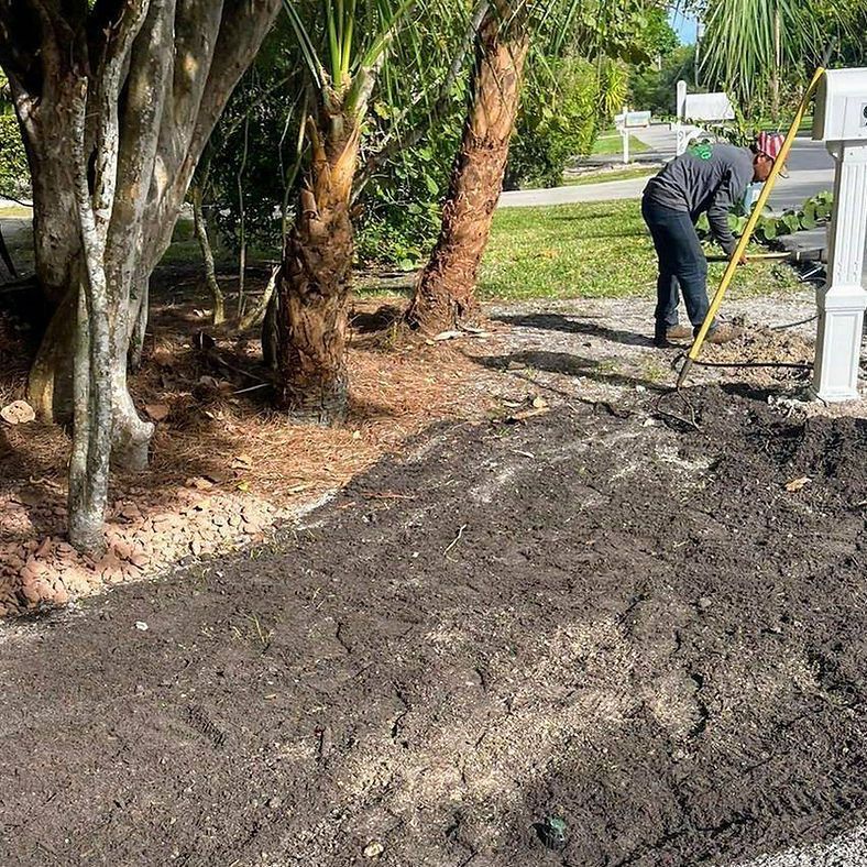 A person rakes dirt near a mailbox and trees in a residential area. There is a large pile of dirt in the foreground.