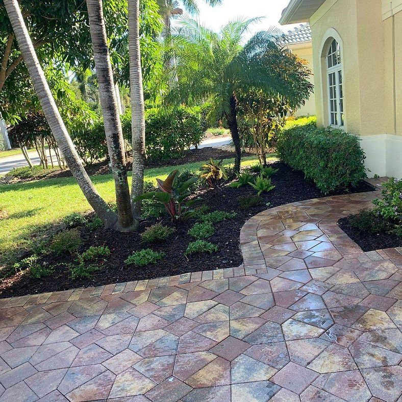 Brick pathway leading to a house with tropical landscaping and palm trees. Ground cover, bushes, and mulch border the path.
